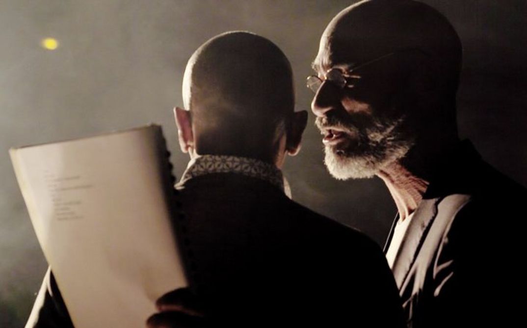 Eine Nahaufnahme von zwei Männern. Der erste steht mit dem Rücken zur Kamera, der zweite ist im linken Seitenprofil zu sehen. Es ist dunkel und neblig. Sie werden von links mit einer Lampe beleuchtet.