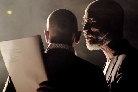 A close-up of two men. The first is standing with his back to the camera, the second can be seen in left side profile. It is dark and foggy. They are illuminated from the left.