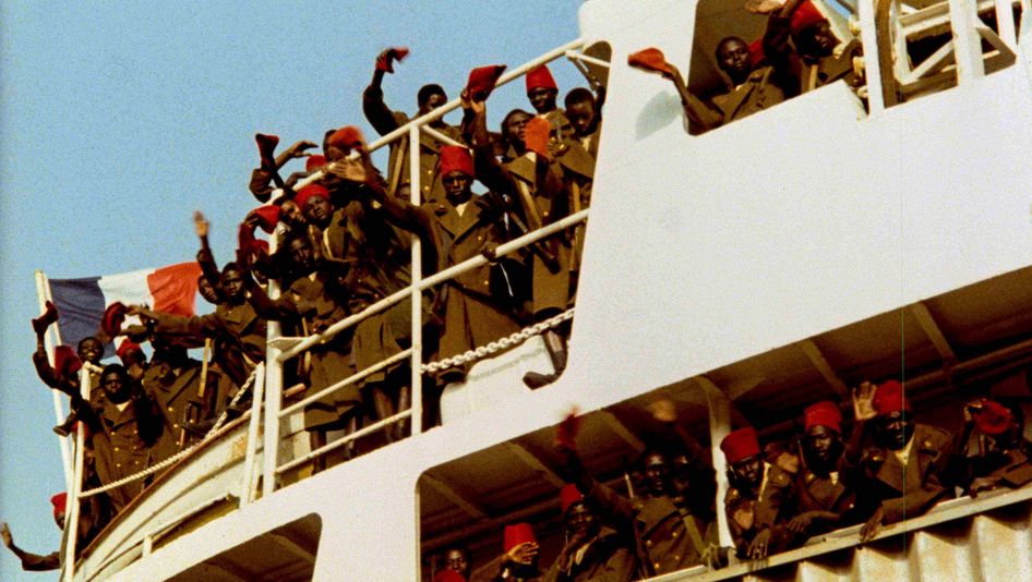 Filmstill from CAMP DE THIAROYE: People in long coats and red caps stand on a ship and wave. The French flag can be seen.