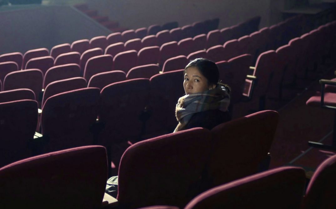 Still from the film "Akyn (Poet)" by Darezhan Omirbayev. A woman sitting alone in an empty theatre is turned and facing the back of the room.
