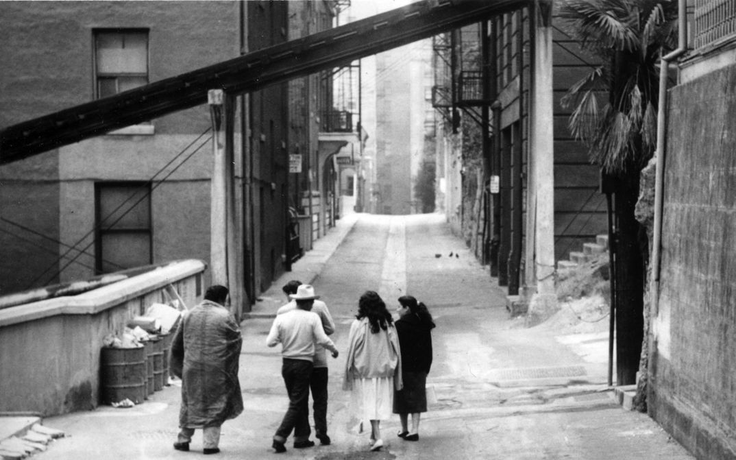 Film still from THE EXILES: A group of people walk along an otherwise empty street.