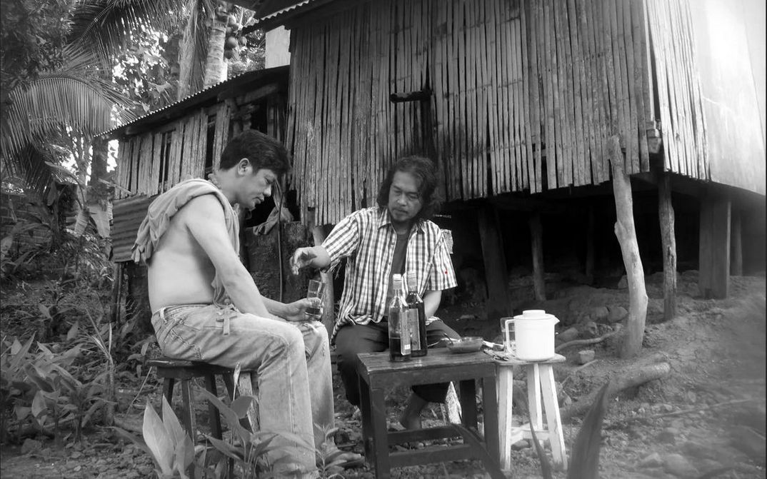 Film still from BUTTERFLIES HAVE NO MEMORIES: Two men are sitting at a small table outside a hut. They have drinks in front of them.