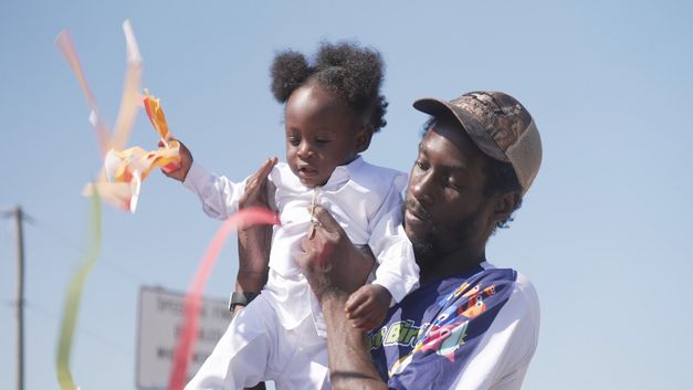 Ein Mann hält Kleinkind im Freien, bunte Bänder flattern im Wind.