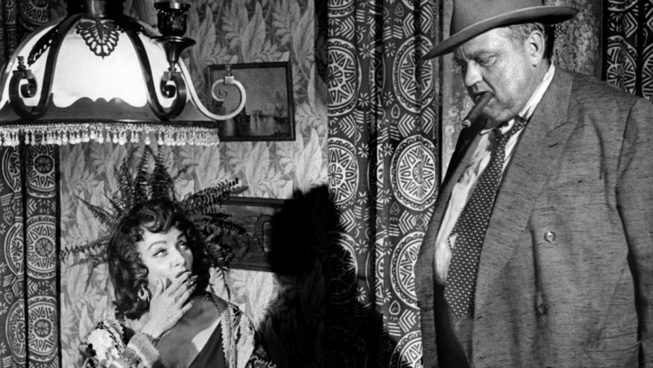 Filmstill from TOUCH OF EVIL: A man with a top hat, a cigar in his mouth and a suit stands next to a seated woman who is also smoking. In the background, patterned wallpaper, curtains and a steelyard.