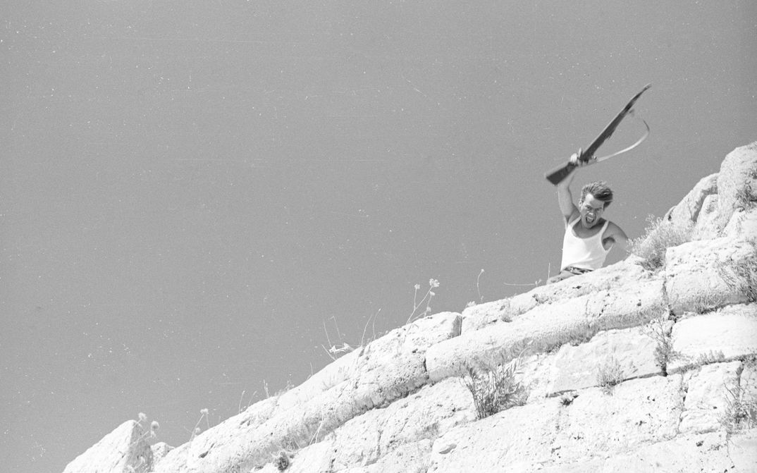 Film still from LEBENSZEICHEN: Above a rocky outcrop, a man is waving a rifle.