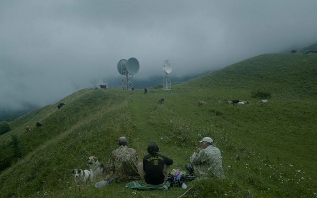 Filmstill aus THE HARVEST. Auf einem Hügel sieht man drei Männer im Gras sitzen. Im Hintergrund grasen Kühe und sind zwei Funktürme mit Satellitenschüsseln zu sehen.