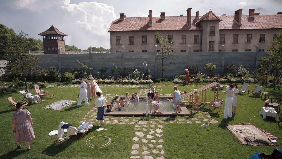 Filmstill aus THE ZONE OF INTEREST: Ein Garten mit einem kleinen Pool ist voller Erwachsener und Kinder, die baden und sich amüsieren. Im Hintergrund sieht man einen Zaun mit Stacheldraht, dahinter ein Wachturm des Konzentrationslagers Auschwitz.