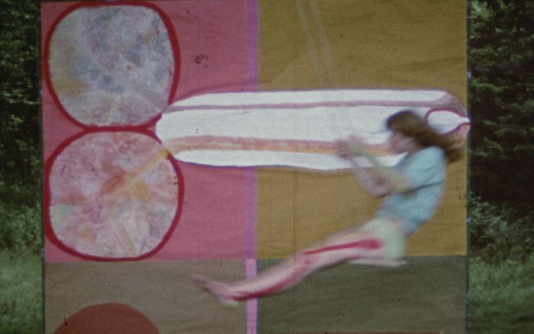 Film still from JÜM JÜM: A young woman swinging. Behind her is a hanging cloth with a colorful pattern.