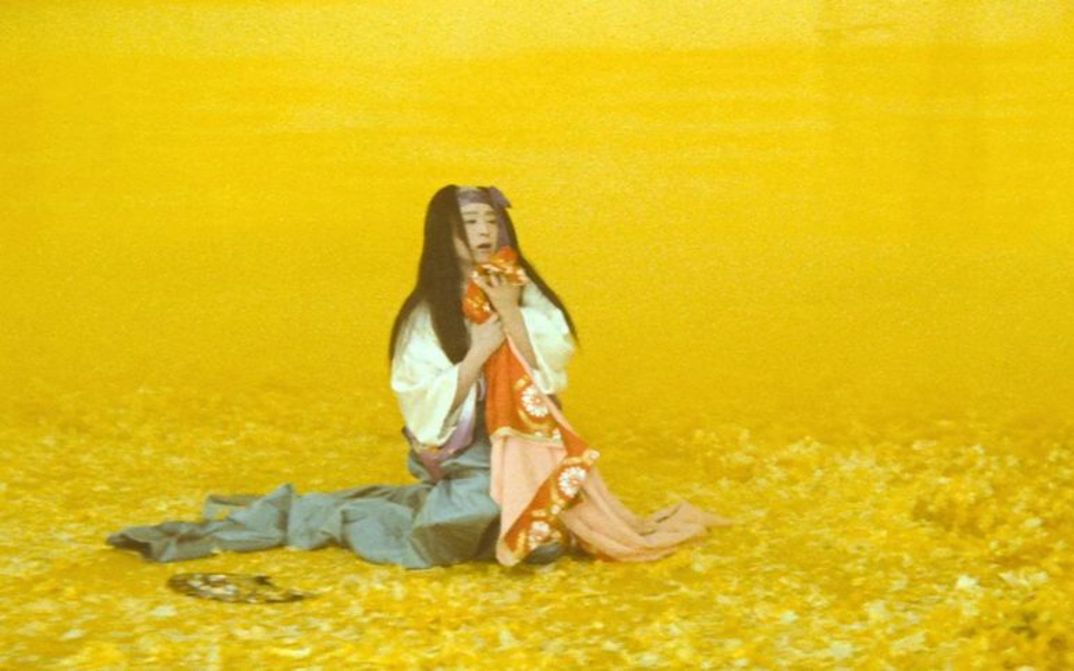 Film still from THE MAD FOX: In a yellow-colored landscape, a person in a kimono kneels and holds something to his chest.