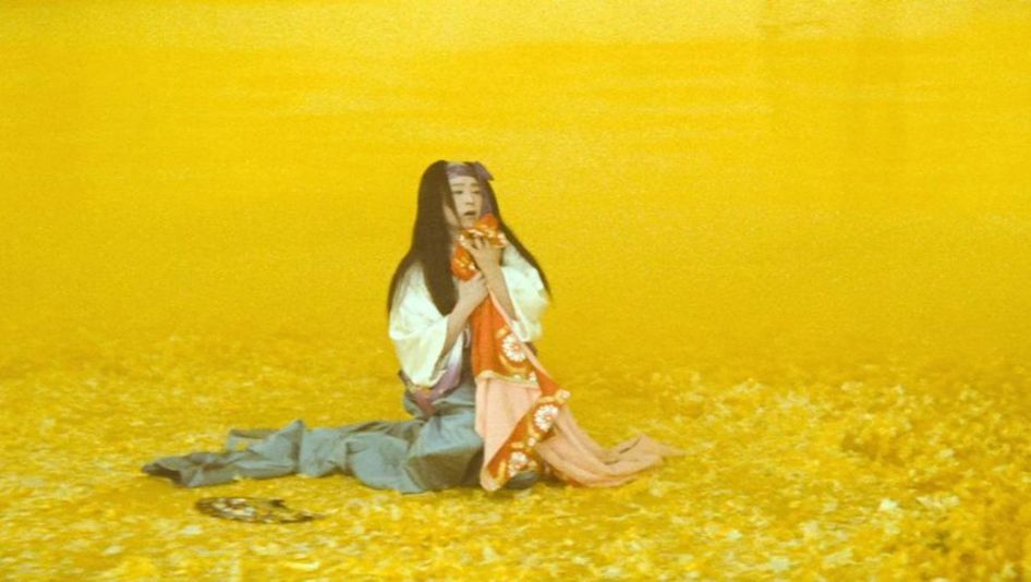 Film still from THE MAD FOX: In a yellow-colored landscape, a person in a kimono kneels and holds something to his chest.