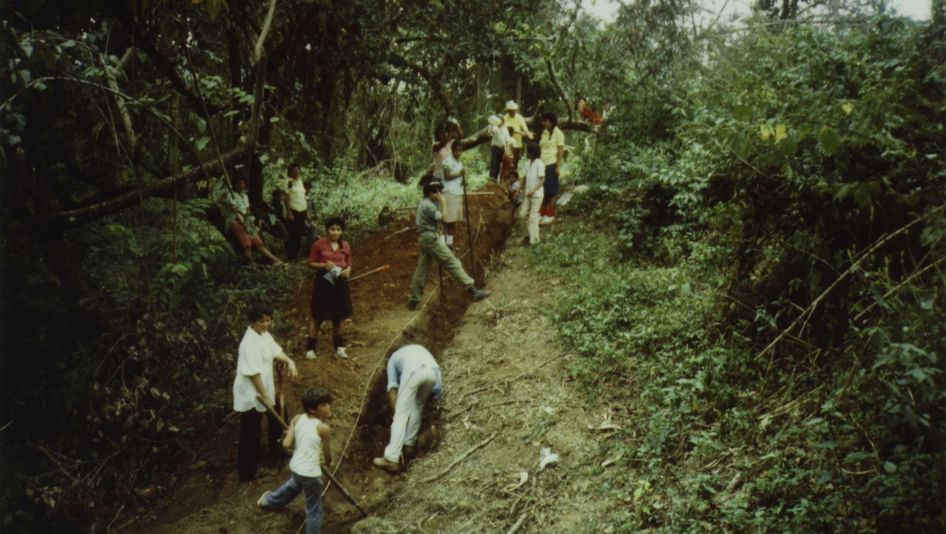 Filmstill from BRIEFE AUS WIWILLI: People dig a furrow in a slope. Around them is forest.