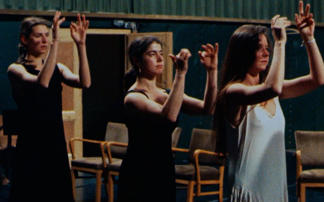 Film still from Was tun Pina Bausch und ihre Tänzer in Wuppertal?: Three women stand one behind the other on a stage with their arms raised.