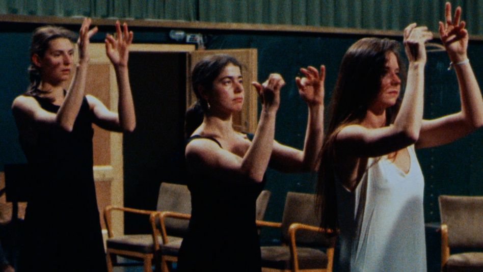 Film still from Was tun Pina Bausch und ihre Tänzer in Wuppertal?: Three women stand one behind the other on a stage with their arms raised.