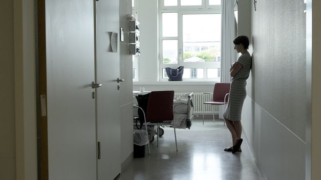 Filmstill aus PALLIATIVSTATION (Palliative Care Unit) von Philipp Döring. Das Bild zeigt eine Frau, die in einem Krankenhauszimmer an der Wand lehnt.