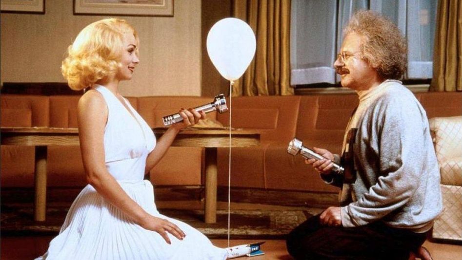 Filmstill aus INSIGNIFICANCE: Zwei Personen, die Marilyn Monroe und Albert Einstein darstellen, knien sich in einem Hotelzimmer gegenüber. Sie beleuchten sich gegenseitig mit Taschenlampen, zwischen ihnen schwebt ein Luftballon.
