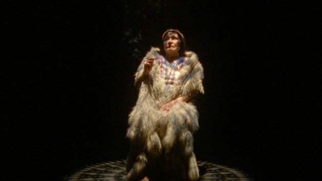 Filmstill aus dem Film „Pidikwe “ von Caroline Monnet. Eine reife Frau sitzt in einem Lichtkreis, in traditioneller indianischer Kleidung. Die Zigarette, die sie zwischen den Fingern hält, gibt Rauch ab, der sich mit der nebligen Atmosphäre des sie umgebenden Raums vermischt.