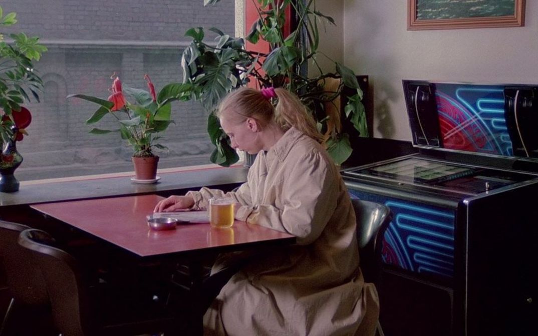 Film still from THE MATCH FACTORY GIRL: A young woman sits alone at an inn table reading a magazine.