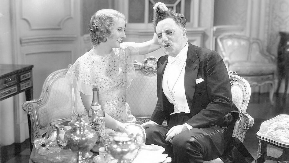 Film still from BABY FACE: A young woman and an older man, both elegantly dressed, sit next to each other on the sofa. She playfully tugs his hair.