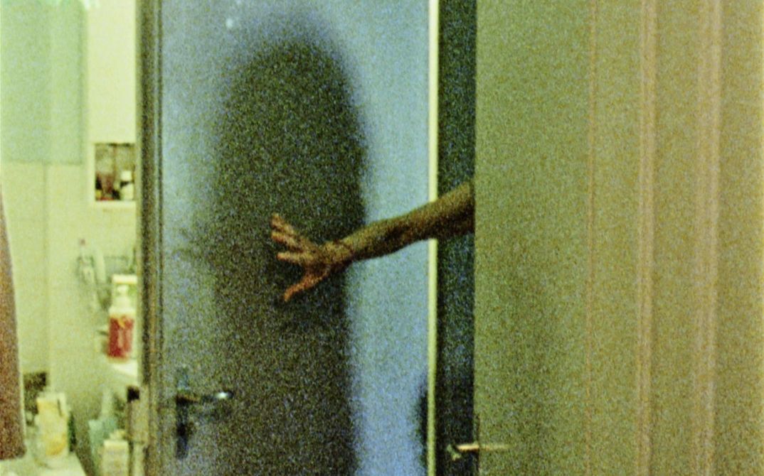 Film still from "Spuren von Bewegung vor dem Eis" by René Frölke. It shows a bathroom. The bathroom door is held open by an arm.