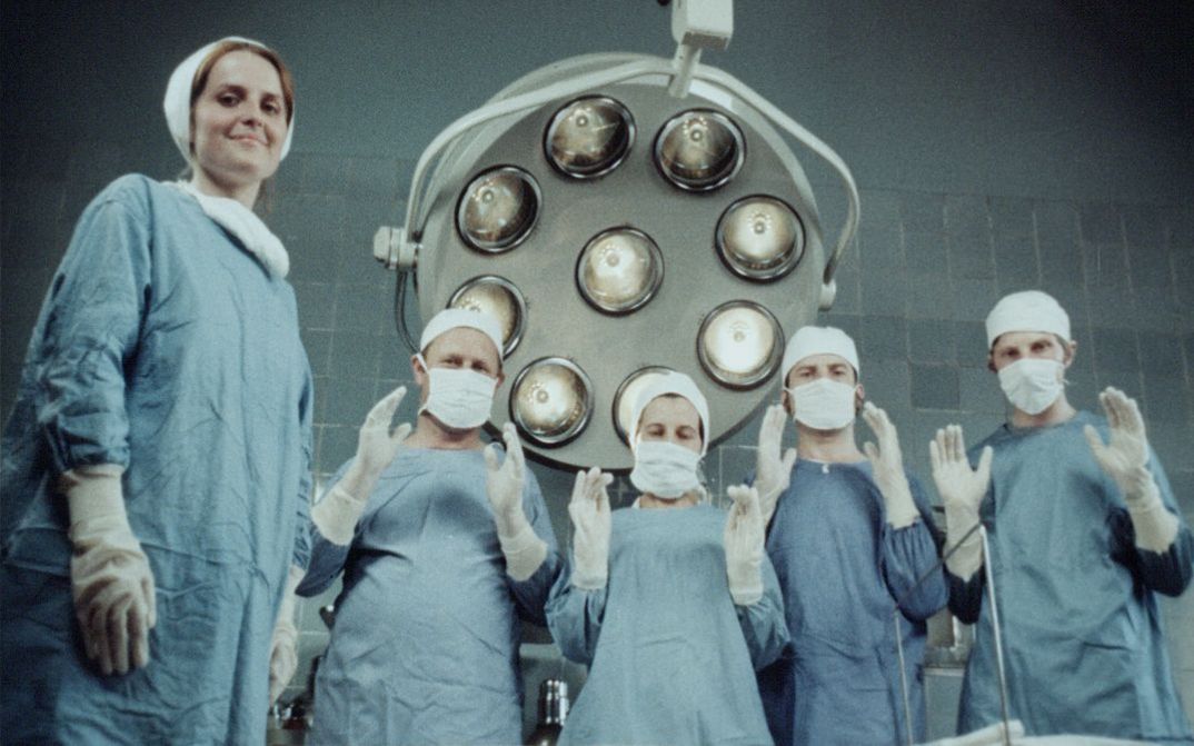 Film still from Zuza Banasińska’s film “Grandmamauntsistercat”. Five nurses wear scrubs and look down at the camera. One of them smiles, whilst the other four, also wearing facemasks, hold both their hands up in front of their chests. A large machine can be seen in the background.