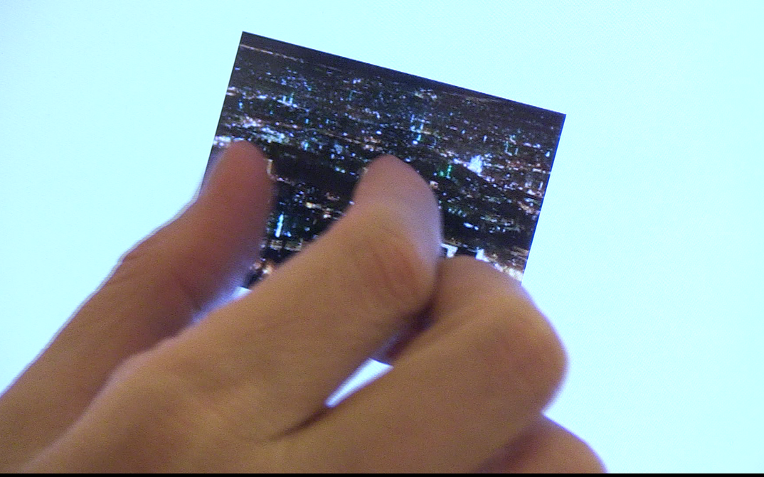 Film still from IN FIELDS OF WORD: CONVERSATIONS WITH SAMAR YAZBEK: A hand holds a picture in front of the sky.