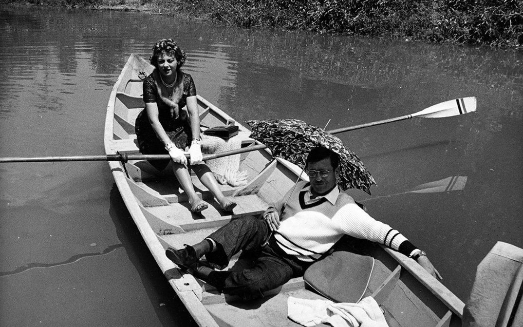 Film still from LA VISITA: A man and a woman are sitting in a rowing boat. She is rowing, he is protecting himself from the sun with an umbrella.