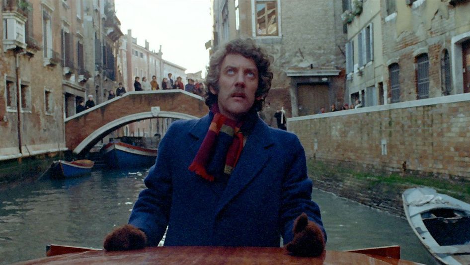 Filmstill aus DON'T LOOK NOW: Ein Mann steht in einem Kanal in Venedig in einer Gondel und blickt nach oben. Im Hintergrund ist eine Brücke mit Menschen zu sehen.