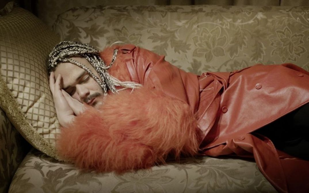 Still from the film „Llamadas desde Moscú“ by Luís Alejandro Yero. A man in a red coat is sleeping on a sofa.