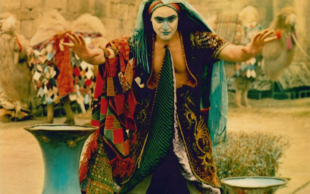 Film still from DIE LEGENDE DER FESTUNG SURAMI: A man in a colorful disguise with a painted face raises his hands imploringly.