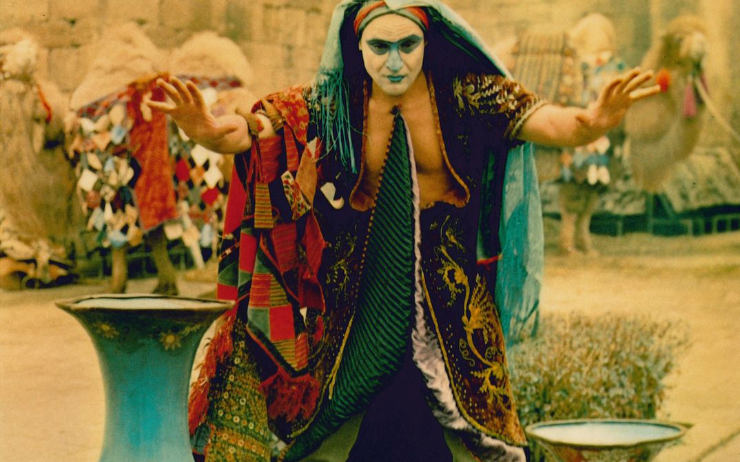 Film still from DIE LEGENDE DER FESTUNG SURAMI: A man in a colorful disguise with a painted face raises his hands imploringly.