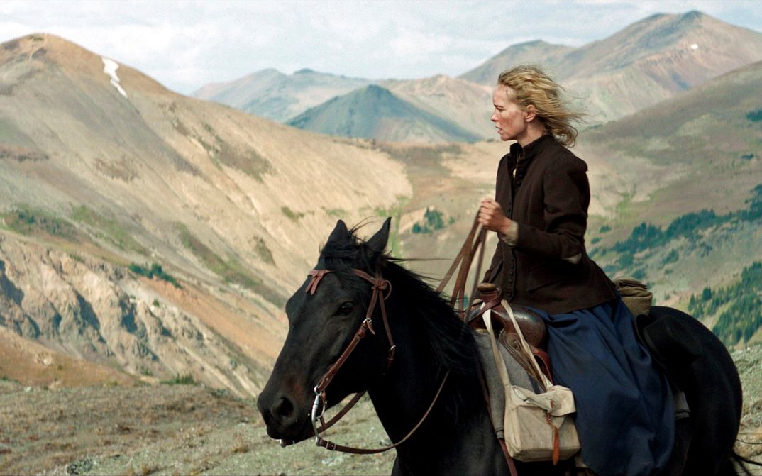 Filmstill from GOLD: A woman sits on a horse and looks into the distance. There are mountains in the background.