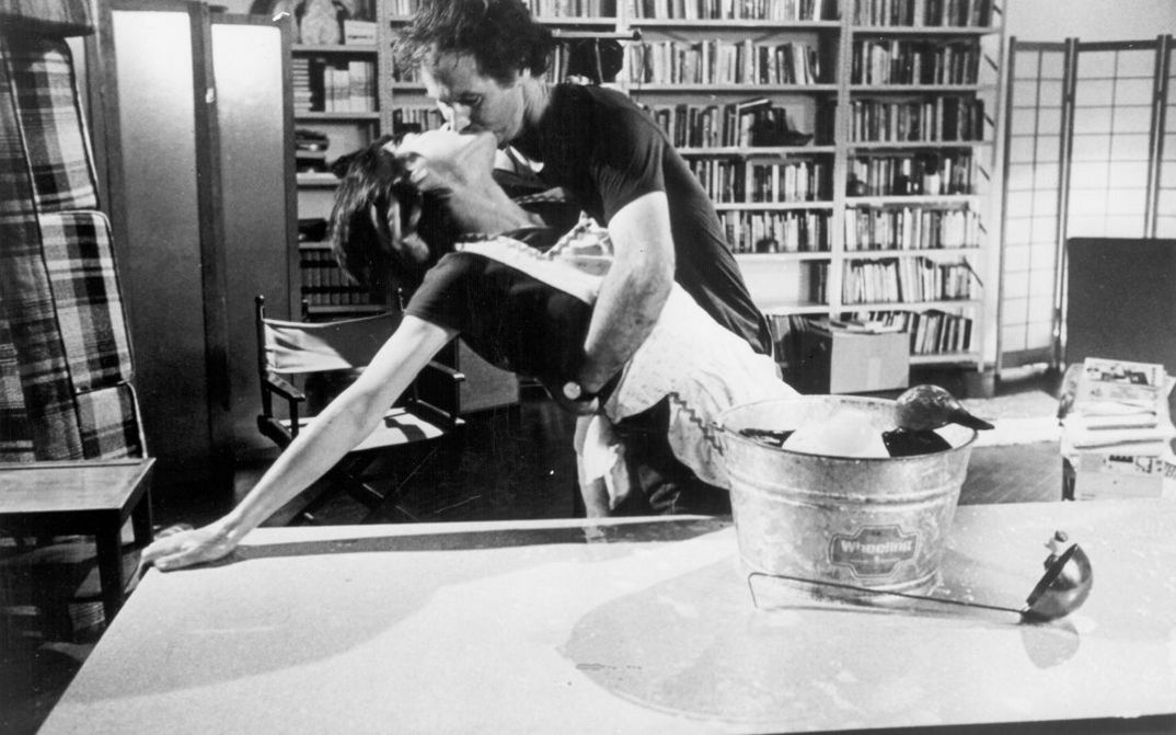 Film still from Yvonne Rainer’s „The Man Who Envied Women“. A women leans on a table, while she kisses a man who is holding her. On the table there is a wooden duck in a bucket and a ladle. In the room in the background there are bookshelves and armchairs.