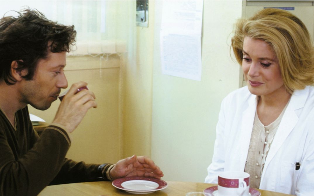 Film still from ROIS ET REINE. Mathieu Amalric and Catherine Deneuve at a table, coffee cups in front of them.