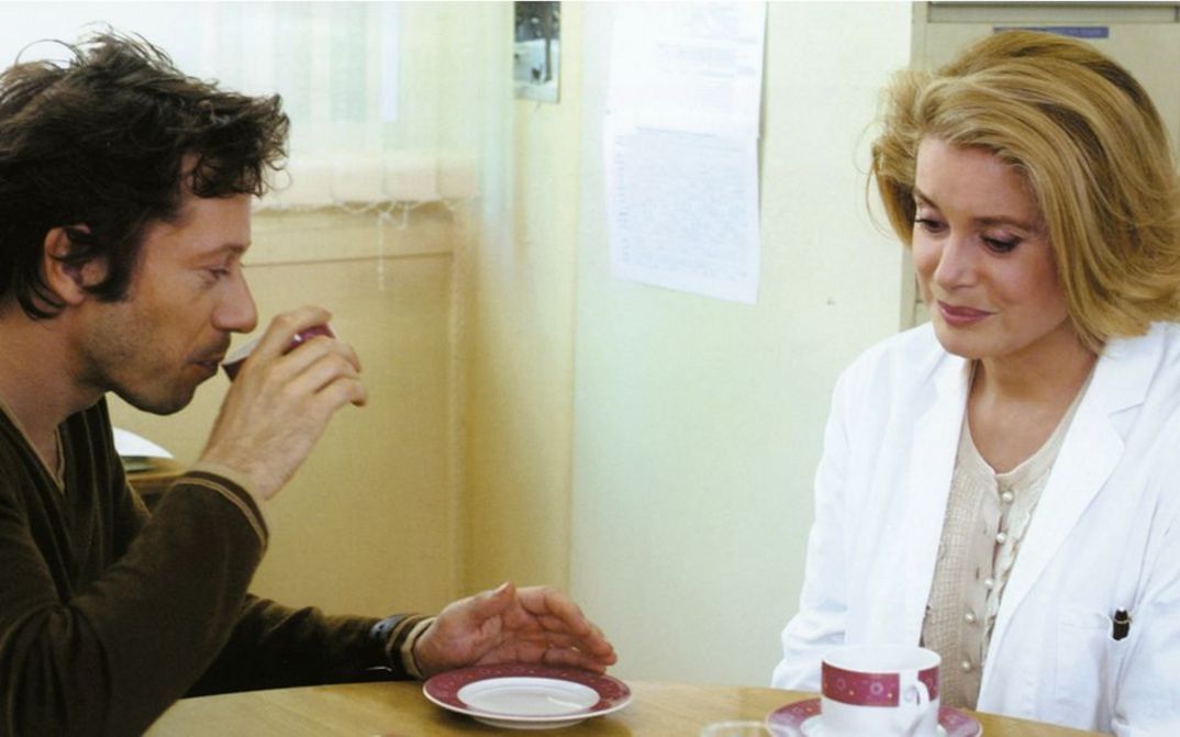 Film still from ROIS ET REINE. Mathieu Amalric and Catherine Deneuve at a table, coffee cups in front of them.
