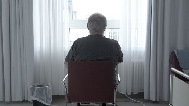 Filmstill aus PALLIATIVSTATION (Palliative Care Unit) von Philipp Döring. Das Bild zeigt eine Person in einem Krankenhauszimmer auf einem Stuhl vor dem Fenster mit dem Rücken zur Kamera.