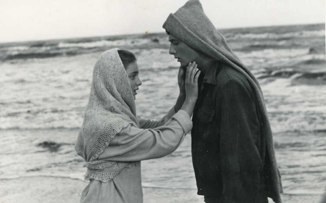 Film still fromm I SEE THE SUN: A girl and a boy stand facing each other in front of the sea, she reaches for his face.
