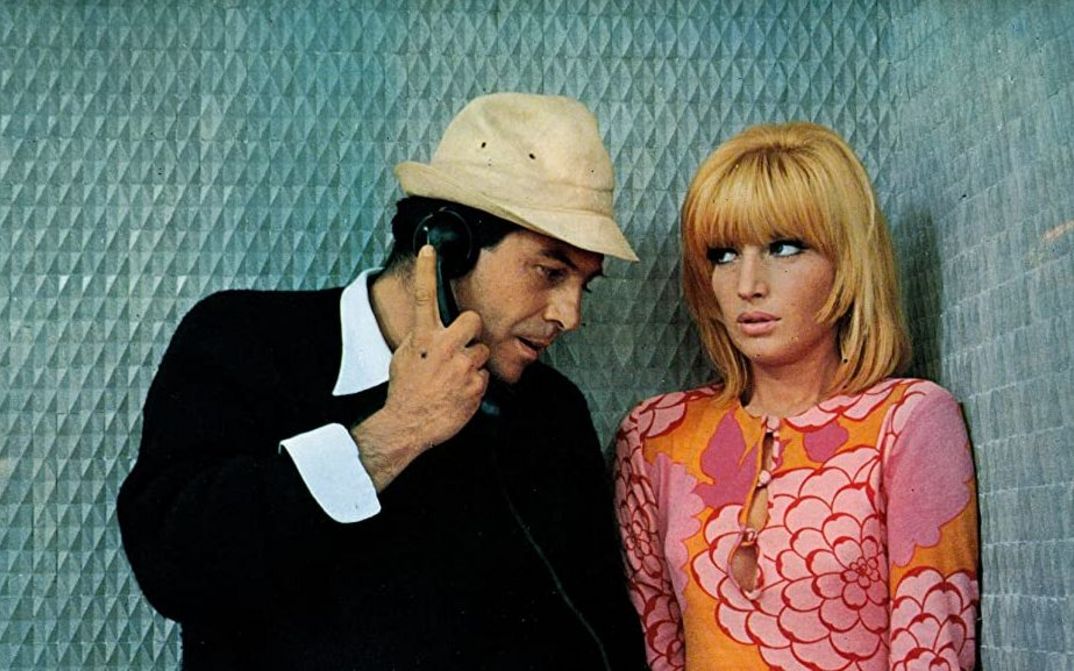 Filmstill aus LE FATE: Ein Mann und eine Frau stehen in einer blau getäfelten Telefonzelle. Er hält den Telefonhörer in der Hand. Sie trägt ein auffällig gemustertes Kleid in orange-rosa.