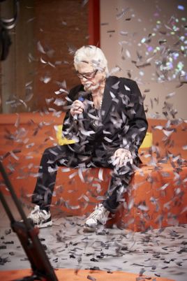 Singer Ricky Shayne sits on an orange sofa with a microphone in his hand. In the foreground, shiny silver strips of confetti swirl through the air.