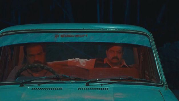 Film still from VAGHACHIPANI by Natesh Hegde. A view through the windshield shows two men in a car. 