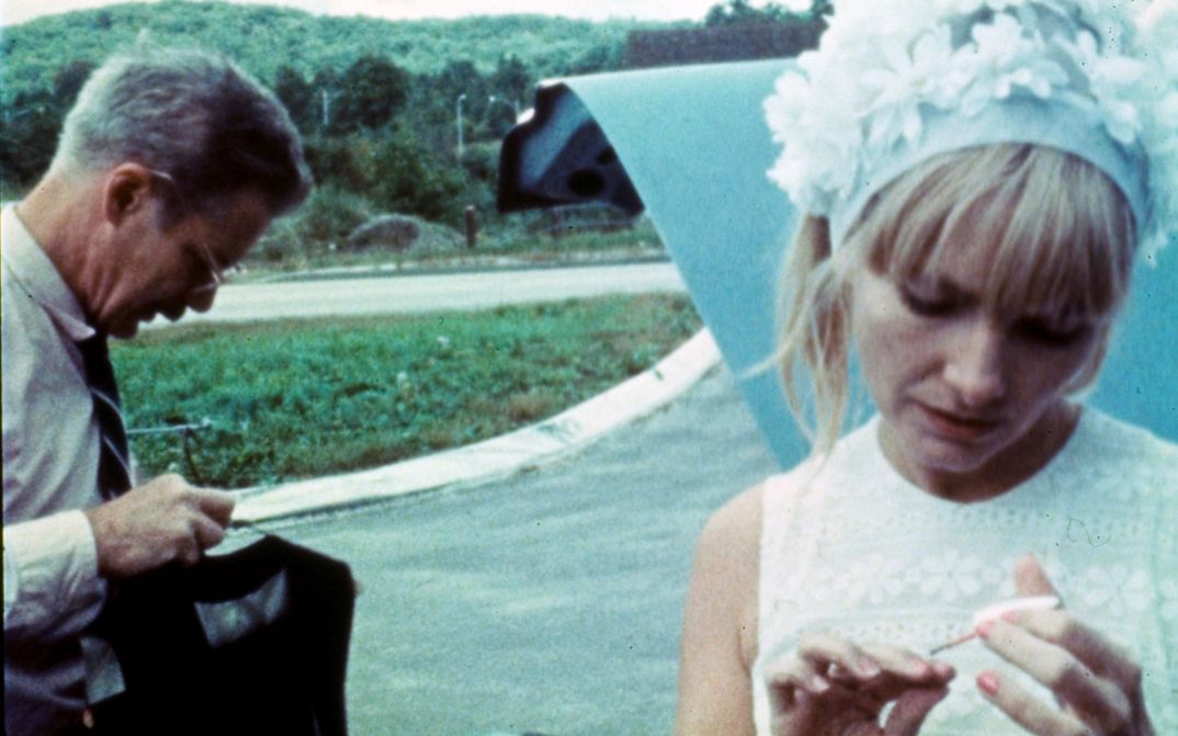 Filmstill aus WANDA: Eine Frau mit geblümter Haube lackiert ihre Nägel. Ein Mann steht neben ihr. Im Hintergrund ist eine Straße.