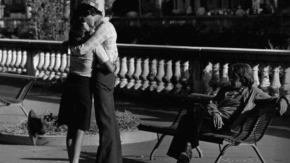 Film still from MES PETITES AMOUREUSES: A girl and a boy are standing close together in a park and kissing. To the right, a boy sits casually on a park bench and watches them.