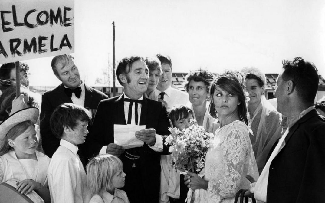 Film still from BELLO, ONESTO, EMIGRATO AUSTRALIA, SPOSEREBBE COMPAESANA ILLIBATA: A group of people surround a bride and groom, someone is holding a sign saying "Welcome Carmela".