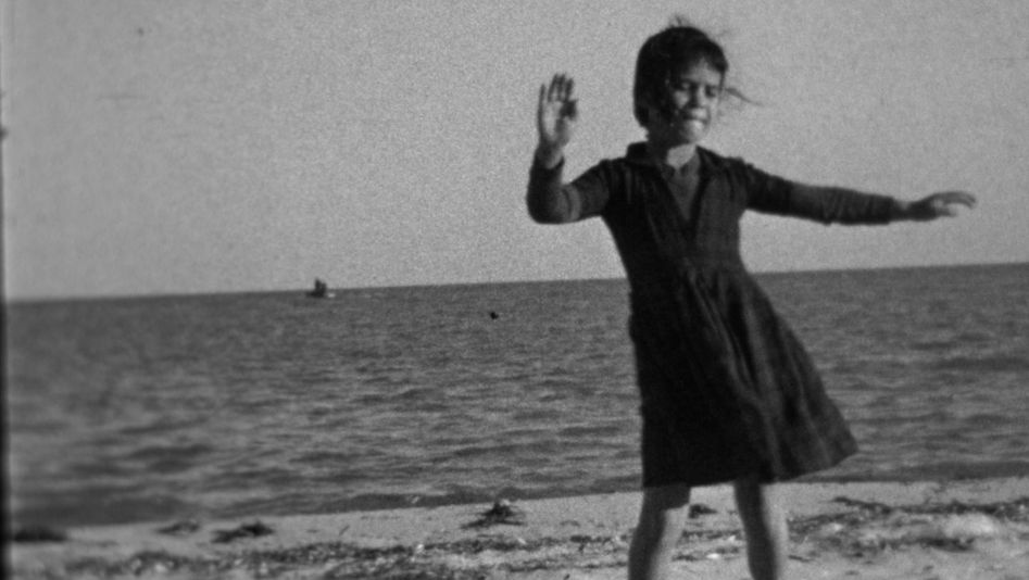 Film still from BALLADE AUX SOURCES: A girl in a dress dancing on the beach, the sea behind her. 