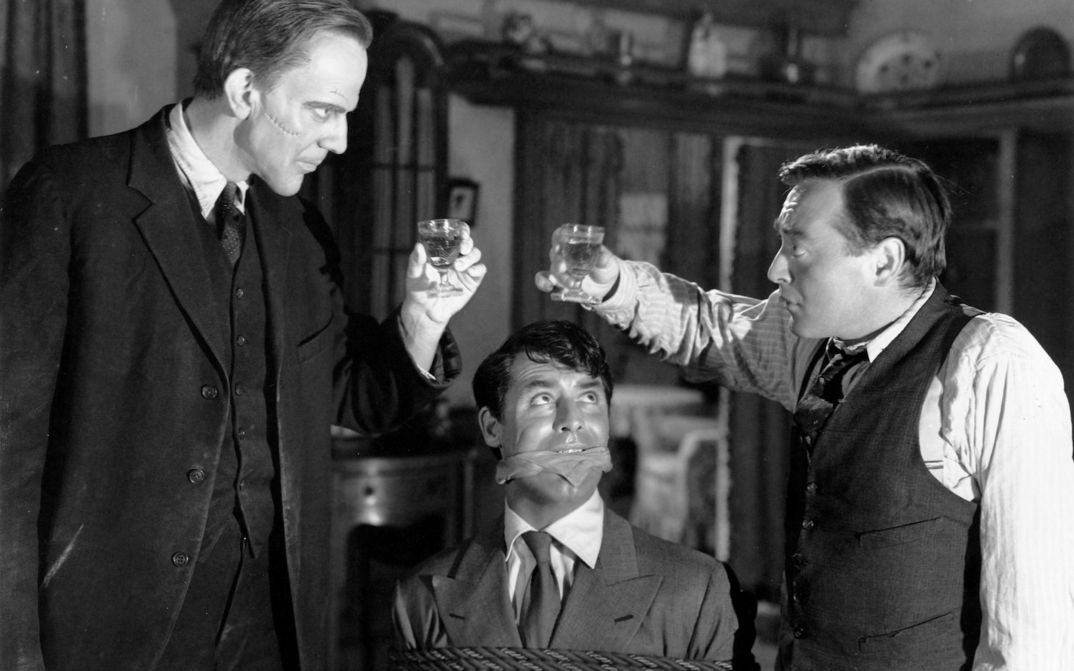 Film still from ARSENIC AND OLD LACE: Two men toast each other, one has a sinister-looking scar on his cheek. A bound and gagged man sits between them and looks up at the two of them.