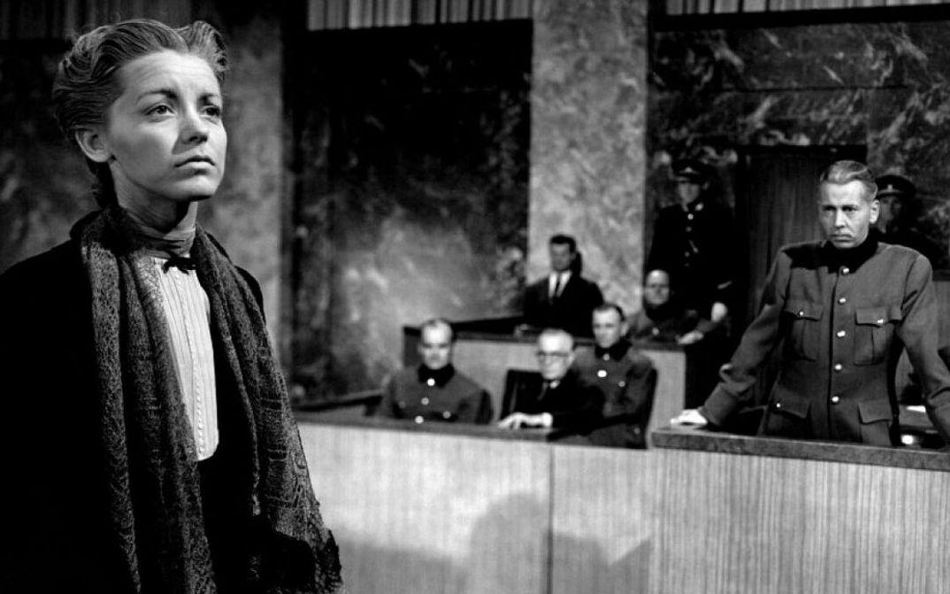 Film still from NONE SHALL ESCAPE: A woman stands in front of the court with her face raised, other people are in the background.