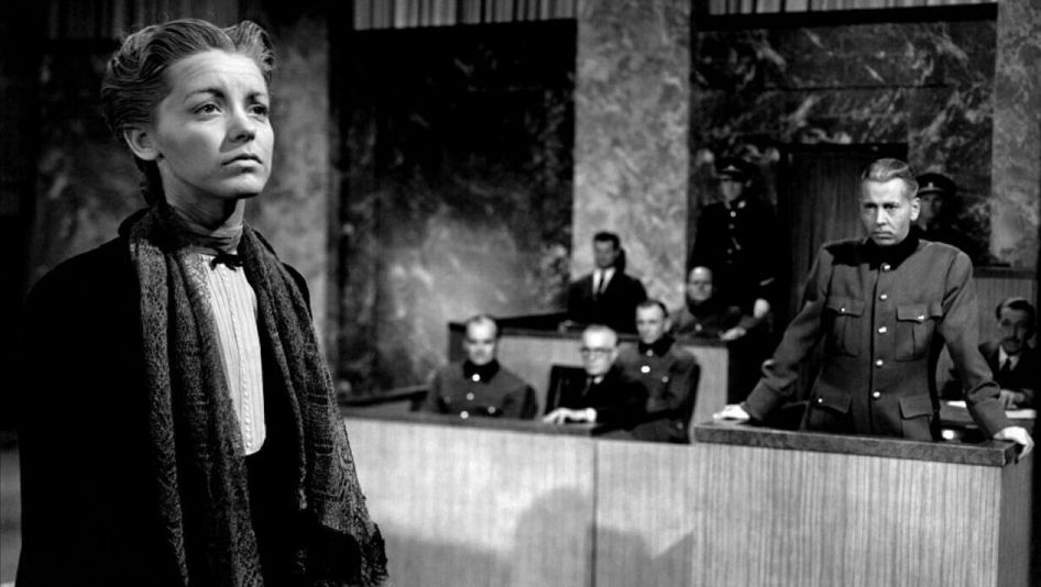 Film still from NONE SHALL ESCAPE: A woman stands in front of the court with her face raised, other people are in the background.