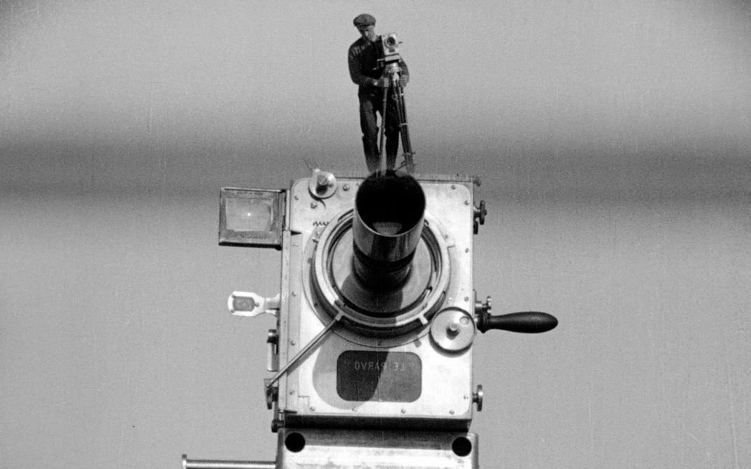 Filmstill from MANN MIT DER KAMERA: A large old camera on which a man is standing with the same camera. He looks like a miniature.