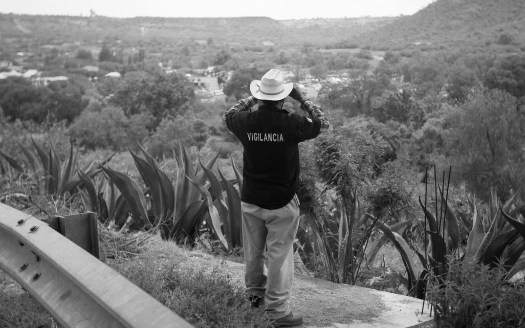Film still from LA EMPRESA: A man looks into the desert with binoculars. He is wearing a T-shirt with the word "Vigilancia" printed on it.