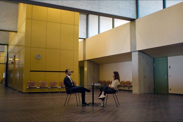 Zwei Personen sitzen sich an einem Tisch in einer großen Halle gegenüber, umgeben von hohen Wänden und leeren Stühlen.