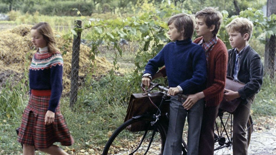 Filmstill aus MES PETITES AMOUREUSES: Ein Mädchen läuft mit einer Schultasche die Straße entlang. Drei Jungen, die an einem Fahrrad stehen, schauen ihr hinterher.